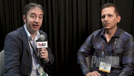 Two men sit side-by-side against a black curtain; one holds a microphone with a logo.