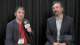 Two men sit side-by-side in front of a black curtain; one holds a microphone.