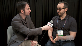 A man in a grey blazer holds a microphone toward a man in a navy polo shirt during an interview.