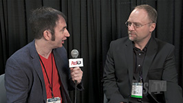 A man in a red shirt and blue blazer holds a microphone while talking to a man in a black suit.