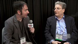 A man in a grey blazer holds a microphone while interviewing a man in a blue shirt and dark blazer.