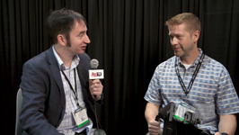 A man in a blazer interviews another man in a plaid shirt against a black curtain.