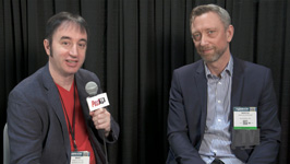 A man in a red shirt and blazer holds a microphone while sitting next to a man in a blue blazer.