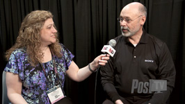 A woman in a floral shirt holds a microphone toward a bald man in a black shirt against a black curtain.