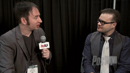 A man in a grey blazer holds a microphone toward a man in a vest and glasses.