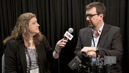 A woman holds a microphone toward a man in a suit during an interview against a black curtain.