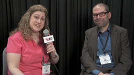 A woman in a pink shirt holds a microphone while a man in a blazer and glasses sits next to her.