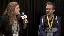 A woman holds a microphone toward a man during an interview against a black curtain background.