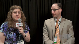 A woman with curly hair speaks into a microphone while a man in a tan suit looks on.