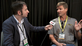 A man in a blazer holds a microphone toward a man in a grey polo shirt during an interview.