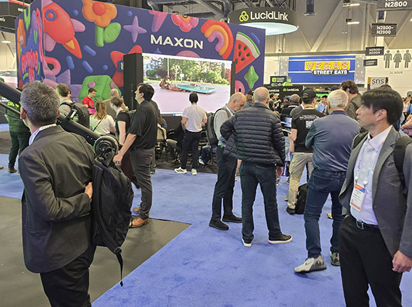 Crowd at a trade show booth with a large screen showing animated graphics and a video. A sign says MAXON.