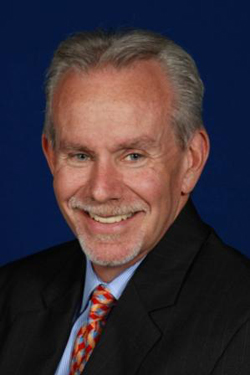 A smiling man with graying hair wearing a dark suit, light blue shirt, and patterned red tie against a blue background.