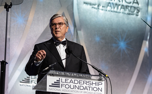 A man in a black tuxedo and glasses speaks at a podium during an awards ceremony.