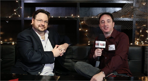 Two men sit on a black leather couch in front of a dark shelf decorated with string lights.
