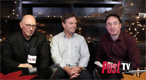Three men sit on a dark couch in front of a shelf decorated with string lights.