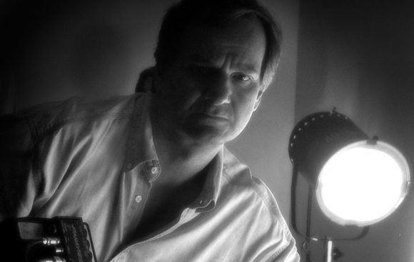 Black and white portrait of a man in a button-down shirt looking at the camera near a bright studio light.