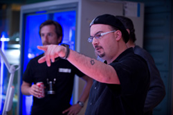 A man with glasses and a black cap points his finger toward something off-camera in a blue-lit room.