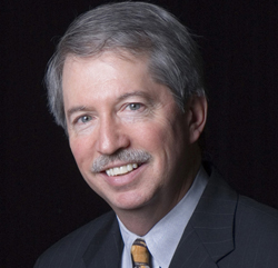 A smiling man with gray hair and a mustache wearing a suit and tie against a black background.