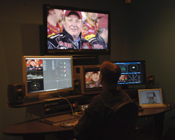 A person sits at a desk viewing several computer monitors and a large television in a dimly lit room.
