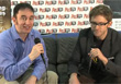 Two men sit on a dark couch holding microphones in front of a branded backdrop.