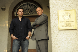 A man in a dark shirt and jeans stands next to a man in a grey suit in front of a police department sign.
