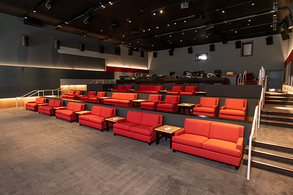 Rows of red sofas arranged on tiered platforms in a dark cinema with overhead lighting.