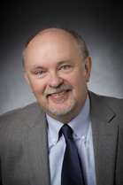 A man with light skin, a bald head, and a grey goatee wearing a grey suit, blue shirt, and dark tie.