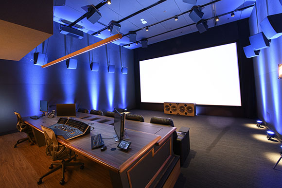 A screening room with a large white screen, a control desk with audio equipment, and multiple speakers mounted on the ceiling
