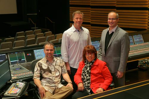 Four people smiling in a recording studio with a large mixing console in the foreground.