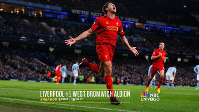 A soccer player in a red kit celebrates with arms outstretched on a grass field in a stadium.