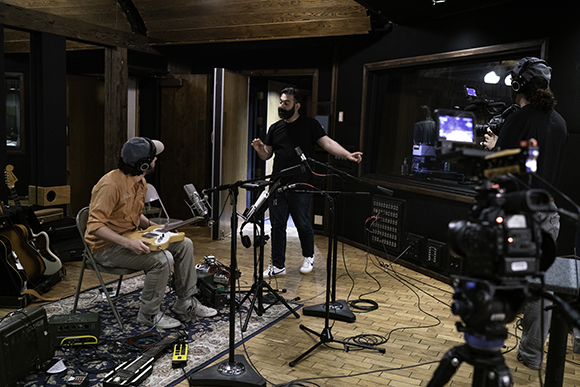 A man sings in a dark recording studio with microphones, cameras, and musical instruments around him.