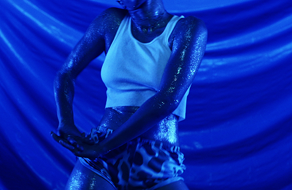 A person with glitter-covered skin wearing a white tank top and patterned shorts under blue light.