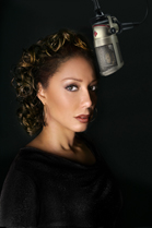 A woman with curly hair looks toward the camera next to a large studio microphone against a black background.