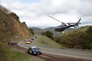 A black helicopter flies over a winding mountain road with cars driving along the curve.