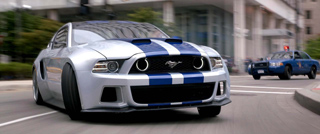 Silver Ford Mustang with blue racing stripes driving through a city street.