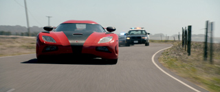 A red and black sports car drives down a paved road with a police car following behind.