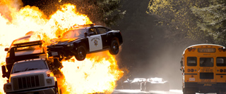 A black police car mid-air during a large explosion next to a white truck and a yellow school bus.