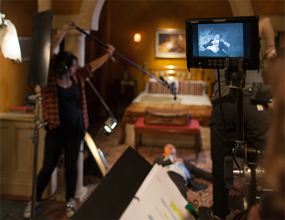 A film set with a person lying on the floor, a boom mic operator, and a monitor displaying the scene.