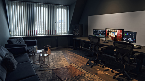 A dark studio with two workstations, large monitors, studio speakers, and a lounge area with a black sofa.