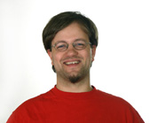 A smiling man with brown hair, glasses, and a goatee wearing a red t-shirt against a white background.