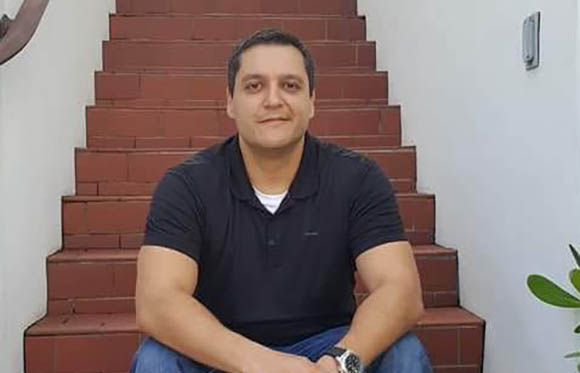 A man in a black polo shirt sitting on red brick stairs against a white wall.