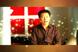 A man in a brown shirt speaks in front of a red window with glowing light spots.