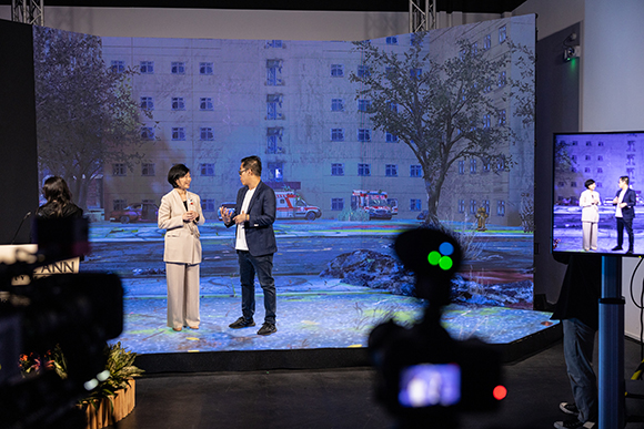 Two people talk in front of a large curved screen showing a building, while cameras film them.