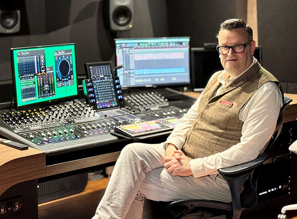 A man in a vest and glasses sits at a large music production mixing desk with multiple monitors.
