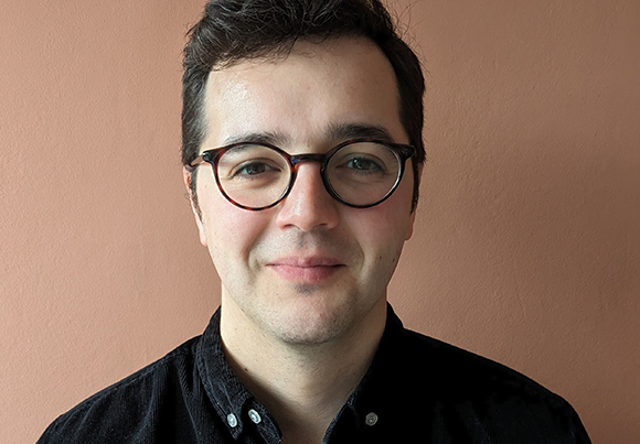 A man with dark hair and round glasses smiling against a solid peach-colored wall.