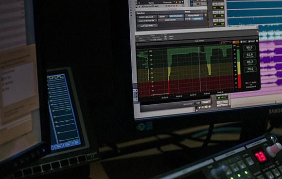 Multiple computer monitors displaying digital audio workstation software with waveforms and frequency graphs.