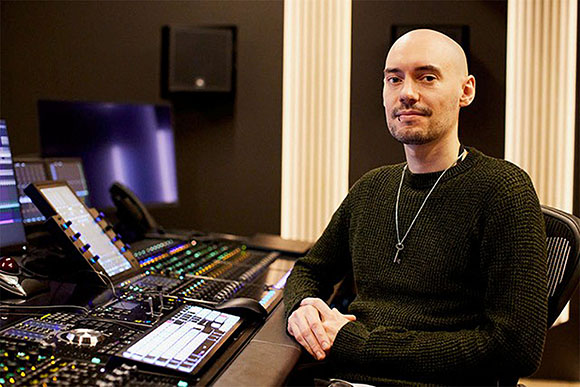 A bald man in a dark green sweater sits behind a large audio mixing desk in a recording studio.