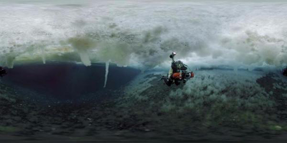 A person in scuba gear swims through dark water beneath a thick, textured layer of white ice.