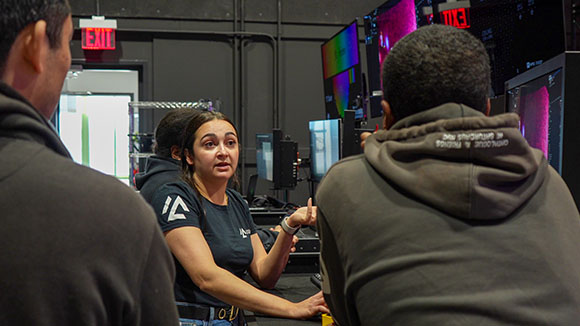 A woman in a dark t-shirt gestures while talking to two men in a room filled with computer monitors.