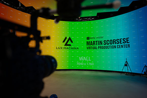 A camera in the foreground films a large, curved LED screen displaying the Lux Machina and NYU logo.